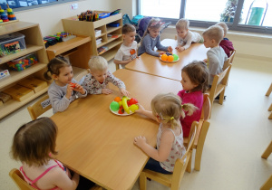 Dzieci siedzą przy stolikach i "częstują się" plastikowymi owocami.