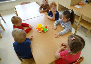 Dzieci siedzą przy stolikach i patrzą na plastikowe owoce postawione na środku.