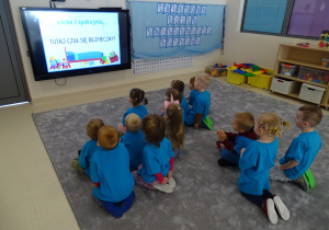 Dzieci siedzą na dywanie i oglądają bajkę na monitorze.