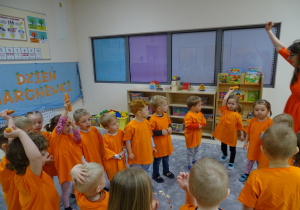 Dzieci z panią stoją w kole ubrane na pomarańczowo. Kilkoro podnosi ręce, w której trzymają marchewki.
