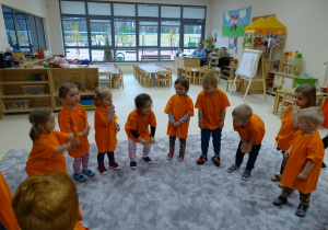 Dzieci z panią stoją w kole ubrane na pomarańczowo. Aniela i Iga trzymają marchewki i z nimi ćwiczą.