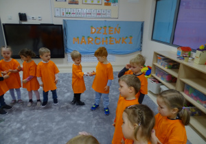 Dzieci z panią stoją w kole ubrane na pomarańczowo. Podają sobie marchewki z ręki do ręki.