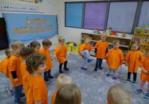 Dzieci z panią stoją w kole ubrane na pomarańczowo. Mikołaj i Wiktor ćwiczą z marchewkami.