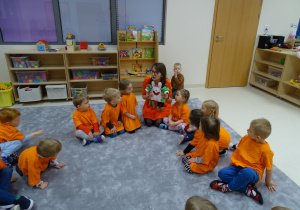 Dzieci siedzą w kole ubrane na pomarańczowo. Pani pokazuje im Treflika i Treflinkę, którzy trzymają królika z papieru.