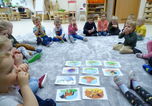 Dzieci siedzą w kole na podłodze wokół rozłożonych obrazków i naśladują zwierzę.