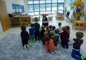 Dzieci siedzą na podłodze i oglądają teatrzyk wystawiany przez nauczycielkę.