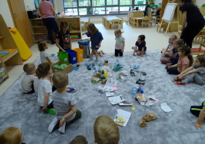 Dzieci siedzą na podłodze, na środku leżą rozrzucone śmieci. Dzieci je sortują.