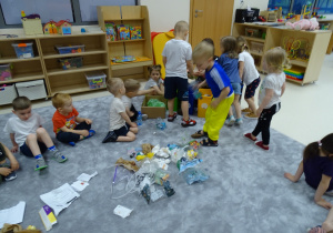 Dzieci siedzą na podłodze, na środku leżą rozrzucone śmieci. Dzieci je sortują.