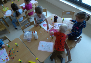 Dzieci siedzą przy stolikach i tworzą portrety dla swoich mam.