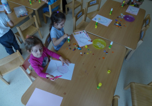 Dzieci siedzą przy stolikach i tworzą portrety dla swoich mam.
