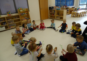 Dzieci siedzą w kole i grają pałeczkami.