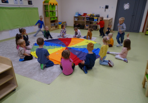 Dzieci siedzą przy kolorowej chuście. Część z nich biega wokół.