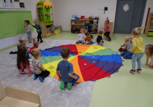 Dzieci siedzą przy kolorowej chuście. Część z nich biega wokół.