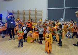 Dzieci w pomarańczowych strojach tańczą i wykonują gesty określone w piosence