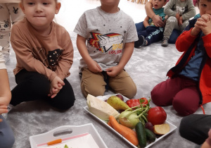 Dzieci siedzą wokół tacek z owocami i warzywami