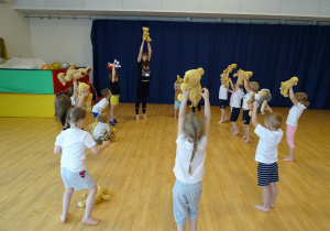 Dzieci ćwiczą na sali gimnastycznej ze swoimi misiami - podnoszą je go góry.