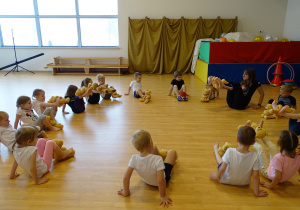 Dzieci ćwiczą na sali gimnastycznej ze swoimi misiami - siad podparty z unoszeniem nóg do góry.