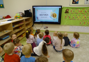Dzieci siedzą na podłodze i oglądają prezentację na tablecie.