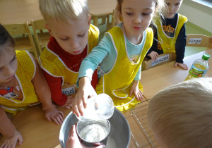 Nikola wsypuje mąkę do misy. Pozostałe dzieci obserwują.