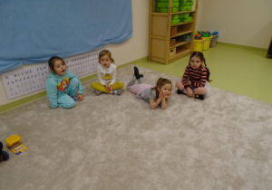 Dzieci siedzą na dywanie po turecku i pokazują emocje za pomocą mimiki.