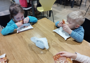 Dzieci siedzą przy stolikach i jedzą pizzę.