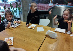 Dzieci siedzą przy stolikach i jedzą pizzę.