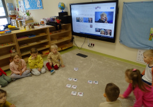 Dzieci siedzą na podłodze i oglądają zdjęcie Wisławy Szymborskiej na ekranie.