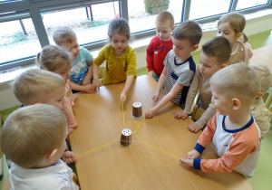 Dzieci zespołowo przenoszą kubki do kawy za pomocą włóczki i gumki.