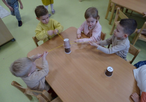 Dzieci zespołowo przenoszą kubki do kawy za pomocą włóczki i gumki.