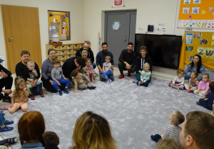 Dzieci wraz z rodzicami siedzą w kole na dywanie