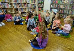 Dzieci siedzą na podłodze pomiędzy regałami książek i podnoszą ręce do góry.