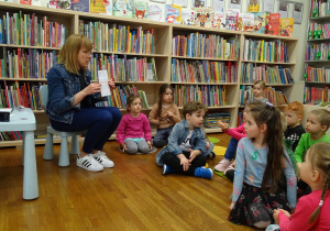 Dzieci siedzą na podłodze pomiędzy regałami książek i słuchają pani bibliotekarki.