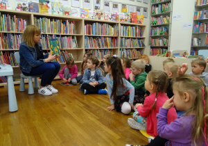 Dzieci siedzą na podłodze pomiędzy regałami książek i słuchają pani bibliotekarki.