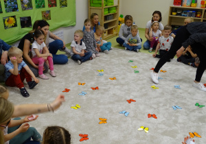 Dzieci z rodzicami siedzą na podłodze, na której porozrzucane są kolorowe motyle.