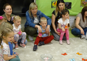 Dzieci z rodzicami siedzą na podłodze i obserwują co się dzieje z pączkami kwiatów umieszczonymi w misce z wodą.