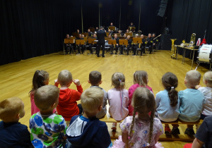Dzieci siedzą i słuchają muzyków grających na instrumentach dętych.