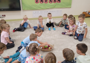 Dzieci siedzą w kole, na środku stoi koszyczek z jabłkami
