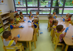 Dzieci siedzą przy stolikach, zakładają jednorazowe rękawiczki