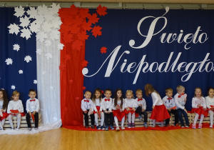 Dzieci siedzą na skrzynkach na tle biało - czerwonej dekoracji.