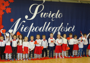 Dzieci stoją na tle biało - czerwonej dekoracji z sercami białymi i czerwonymi w rękach.