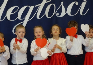Dzieci stoją na tle biało - czerwonej dekoracji z sercami białymi i czerwonymi w rękach.