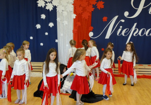 Dziewczynki tańczą w kółku z białymi i czerwonymi wstążkami.