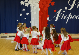 Dziewczynki tańczą w kółku z białymi i czerwonymi wstążkami.