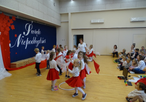 Dzieci tańczą z białymi i czerwonymi chustkami i biało - czerwonymi flagami.