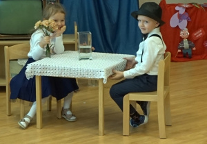 Dziewczynka z kwiatami w ręku siedzi przy stole razem z chłopcem.