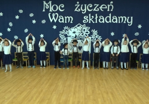 Dzieci spiewają, stoją w rzędzie i pokazują daszki nad głowami.