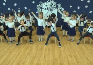 Dzieci tańczą - dziewczynki z goździkami, chłopcy na krzesłach z kapeluszami.