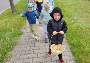 Dzieci w parach idą chodnikiem, chłopiec z przodu trzyma koszyczek