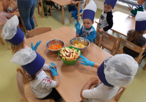 Dzieci siedzą przy stoliku pełnym owoców i marchewki.