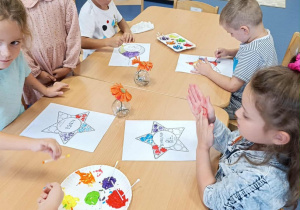 Dzieci przy stoliku ozdabiają wizytówkę patyczkami kosmetycznymi umoczonymi w kolorowej farbie
